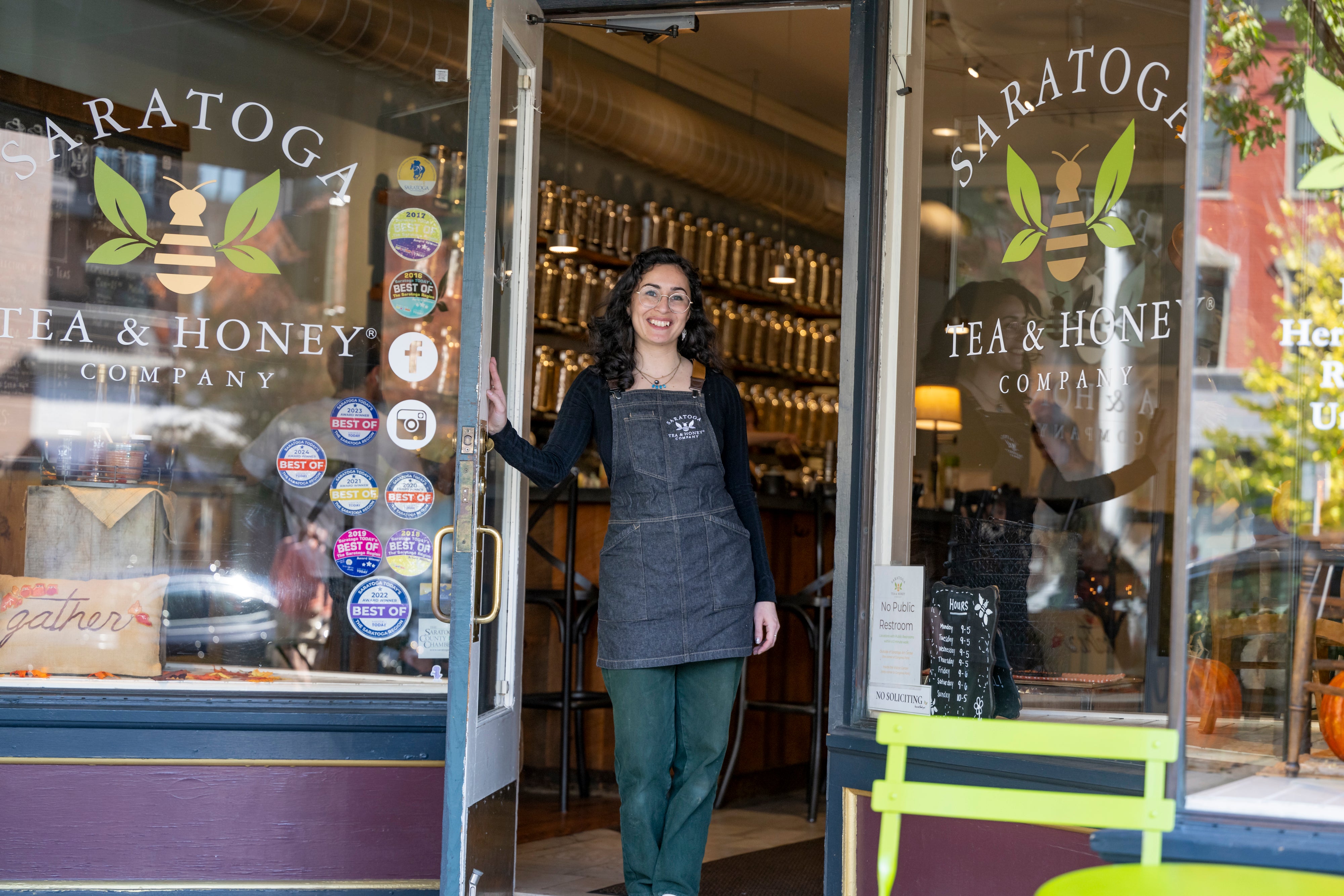 Welcoming scene opening the door at Saratoga Tea & Honey Co. A woman with black hair in a denim apron holds open the door to Saratoga Tea & Honey Co. with a welcoming glow inside.