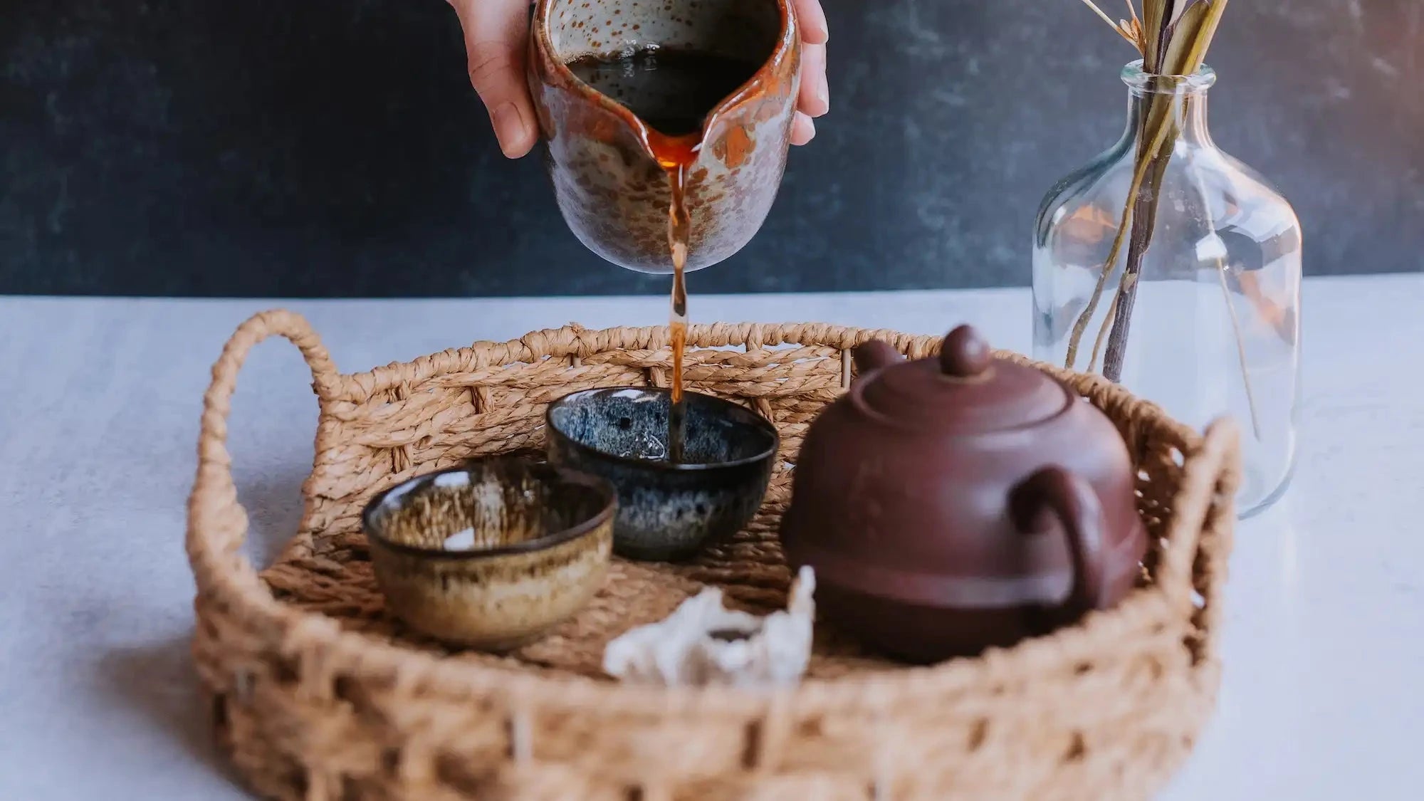 hand pouring brewed pu erh (aged tea) into small cups from sharing pitcher next to yixing clay pot and a small tuocha (nest) cake of aged tea