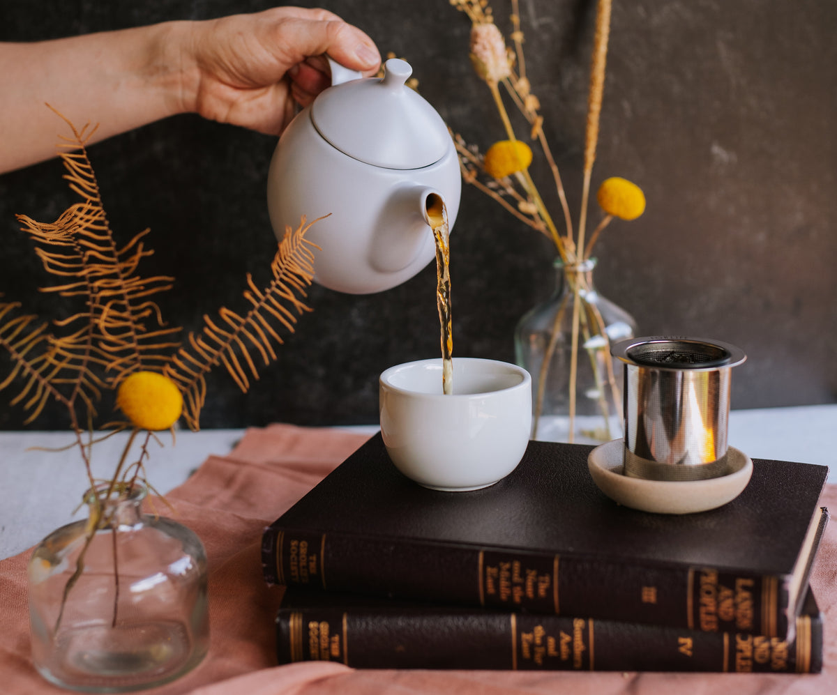 A hand pours tea from a teapot into a cup resting on a stack of old books.