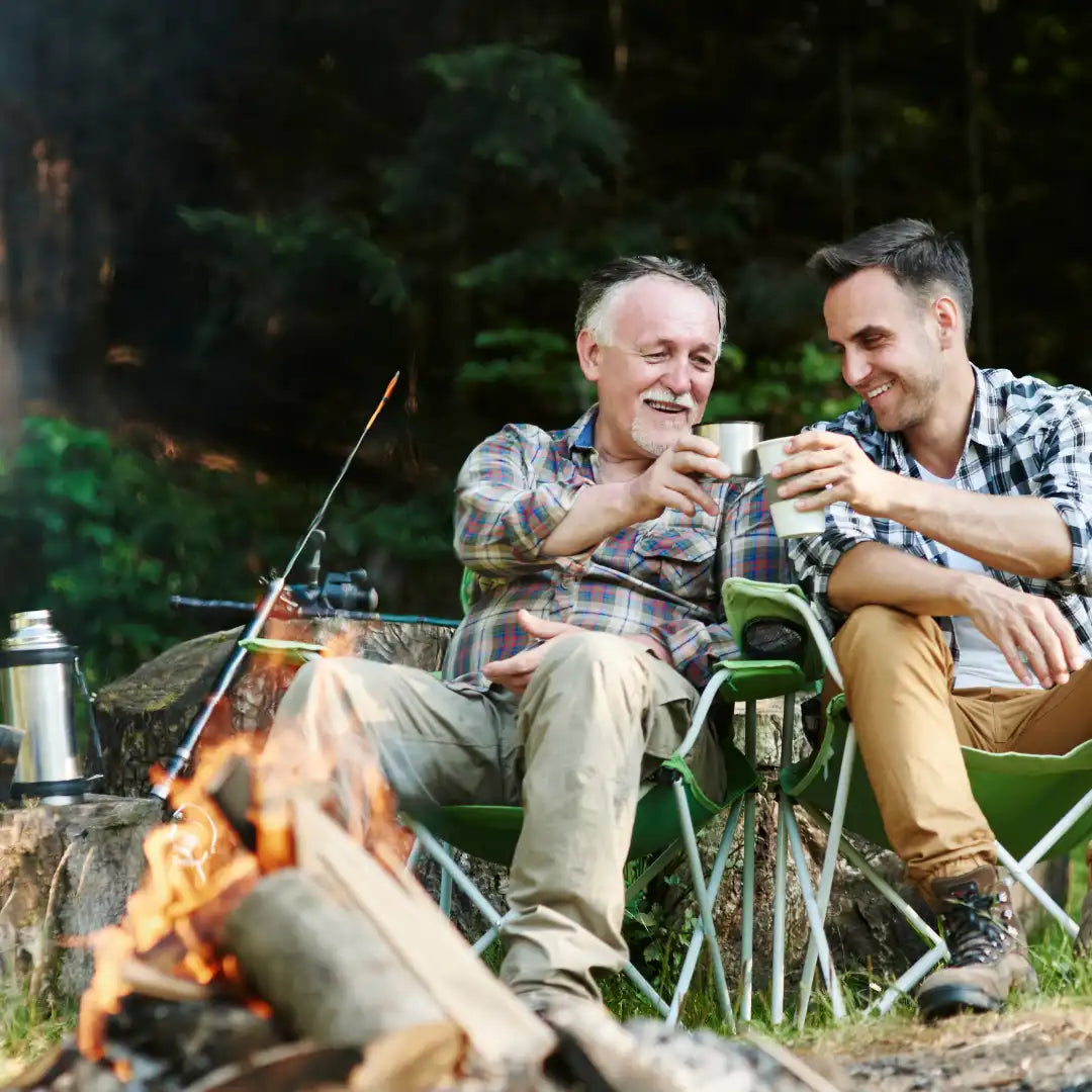 two men sitting at a campfire cheersing with mugs