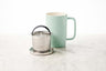 showing three parts of the dew brew in mug: stainless steel lid and basket rest, stainless steel infuser basket, and minty aqua ceramic mug with large handle