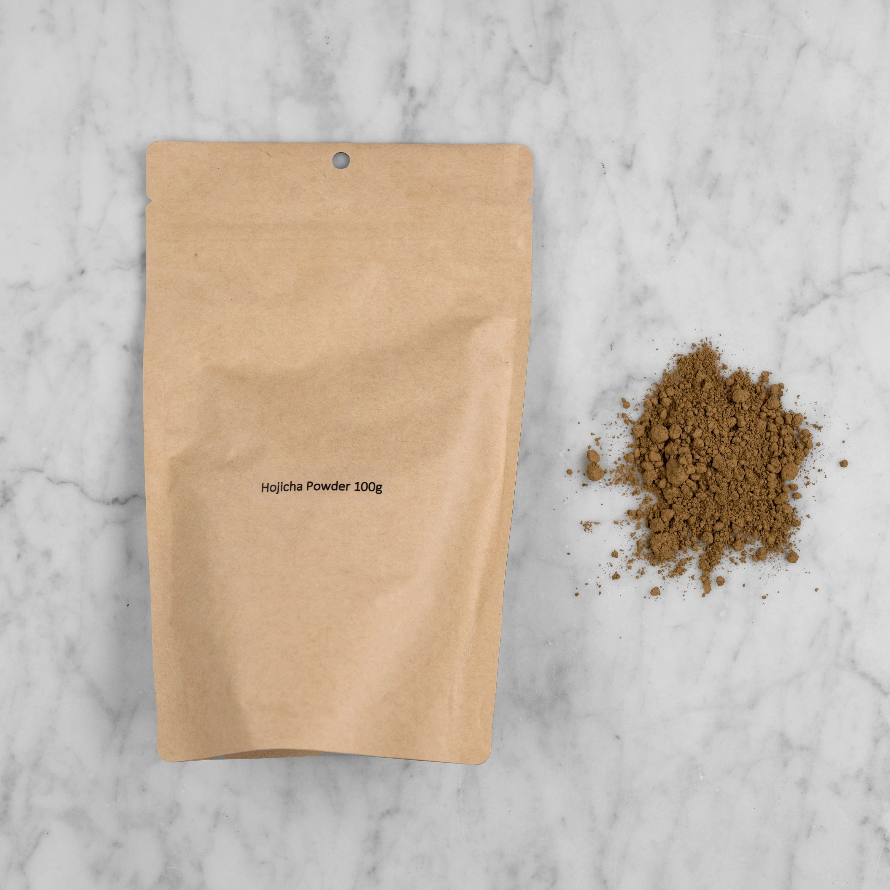 Hojicha Powder packaging and pile of ground tea. The hojicha powder comes in a kraft pouch with a label reading: Hojicha Powder 100g. Next to the pouch is a pile of ground roasted green tea. It appears brown in color and is finely ground. 
