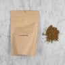 Hojicha Powder packaging and pile of ground tea. The hojicha powder comes in a kraft pouch with a label reading: Hojicha Powder 100g. Next to the pouch is a pile of ground roasted green tea. It appears brown in color and is finely ground. 