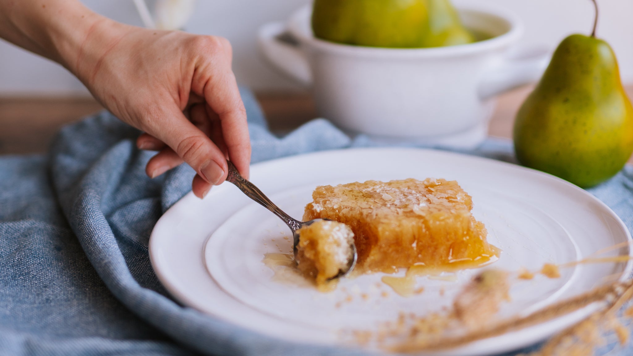Aesthetic scene of honeycomb and fruit. A white woman's hand uses a small silver spoon to scoop a corner off a block of golden, dripping honeycomb on a dinner plate. 