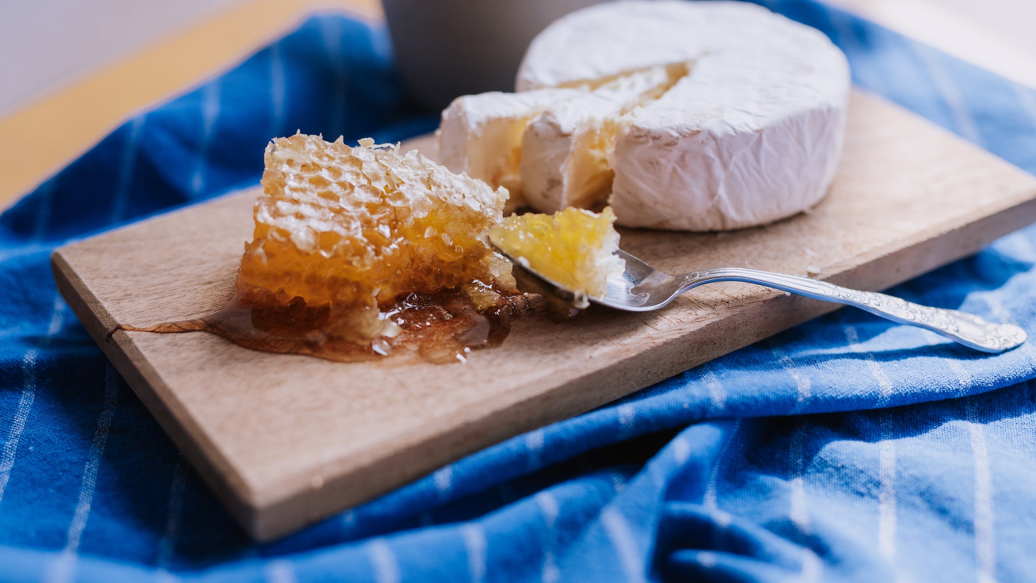 A chunk of golden honeycomb oozing honey sits on a wooden board with a spoon and round wheel of soft cheese with two slices cut out.