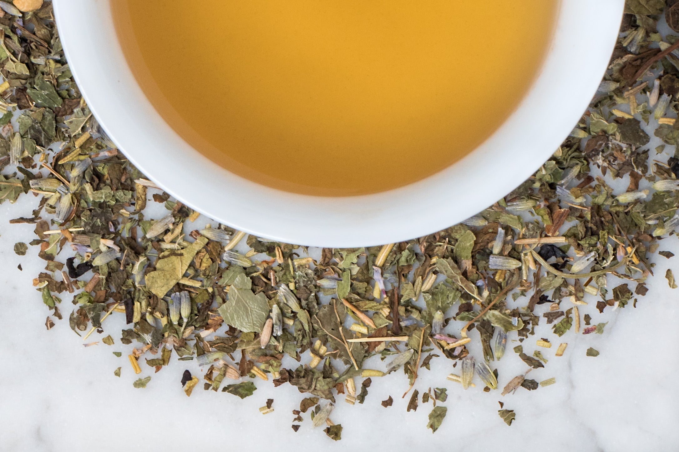 La Provençale leaves and brewed tea. Small, torn khaki-brown and green peppermint, lavender, rosemary, and lemon balm leaves surround a cup of golden-yellow brewed tea.