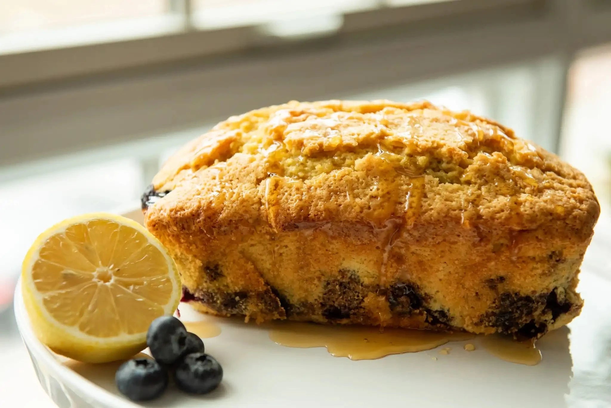 loaf of blueberry ginger lemon bread resting on a white plate with fresh blueberries and a half a lemon, all drizzled with honey