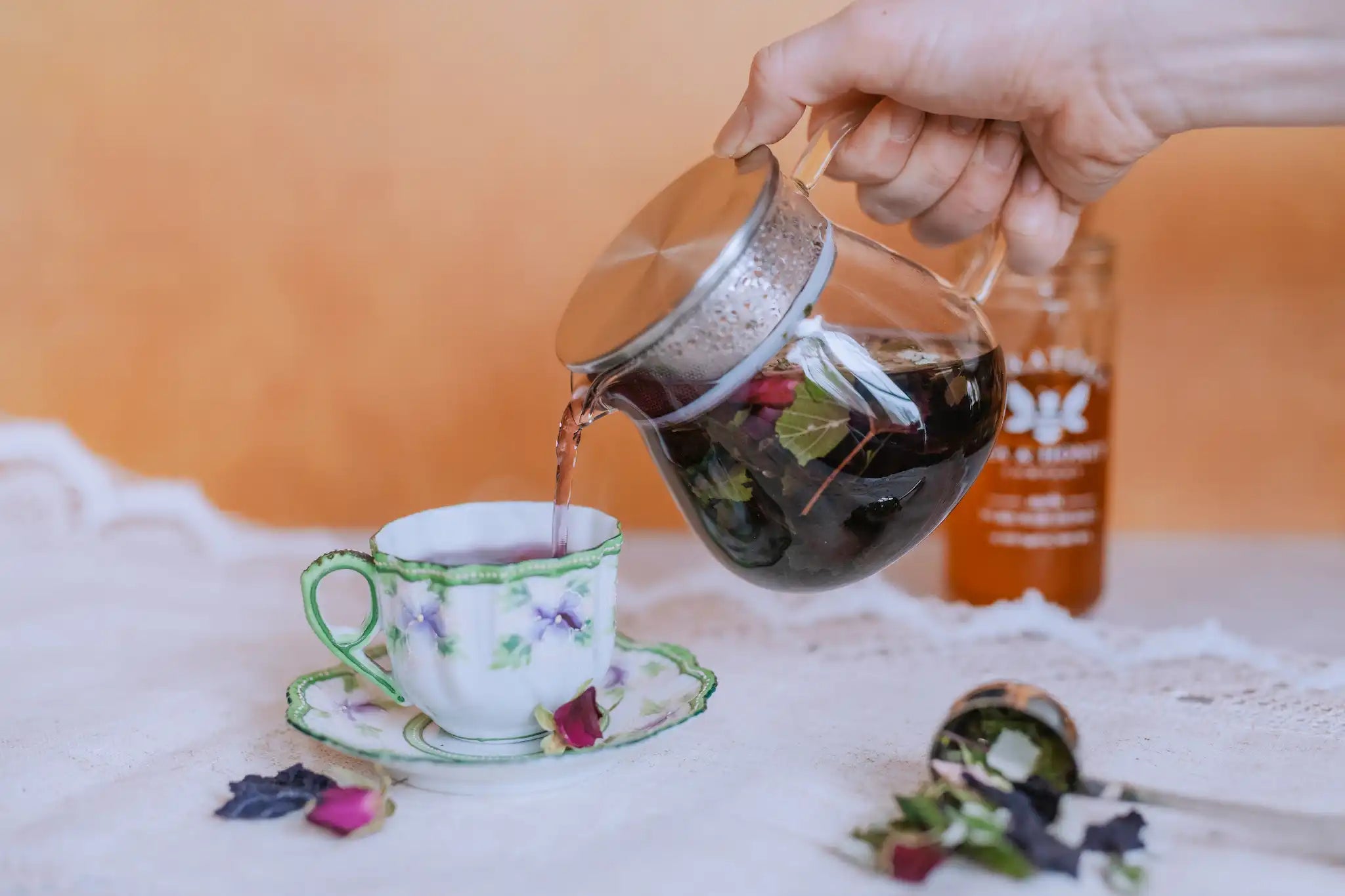 pouring purple herbal tisane la vie en rose from glass vetro teapot from Saratoga Tea & Honey Co into a vintage tea cup with spilled tea in foreground and jar of honey in the background