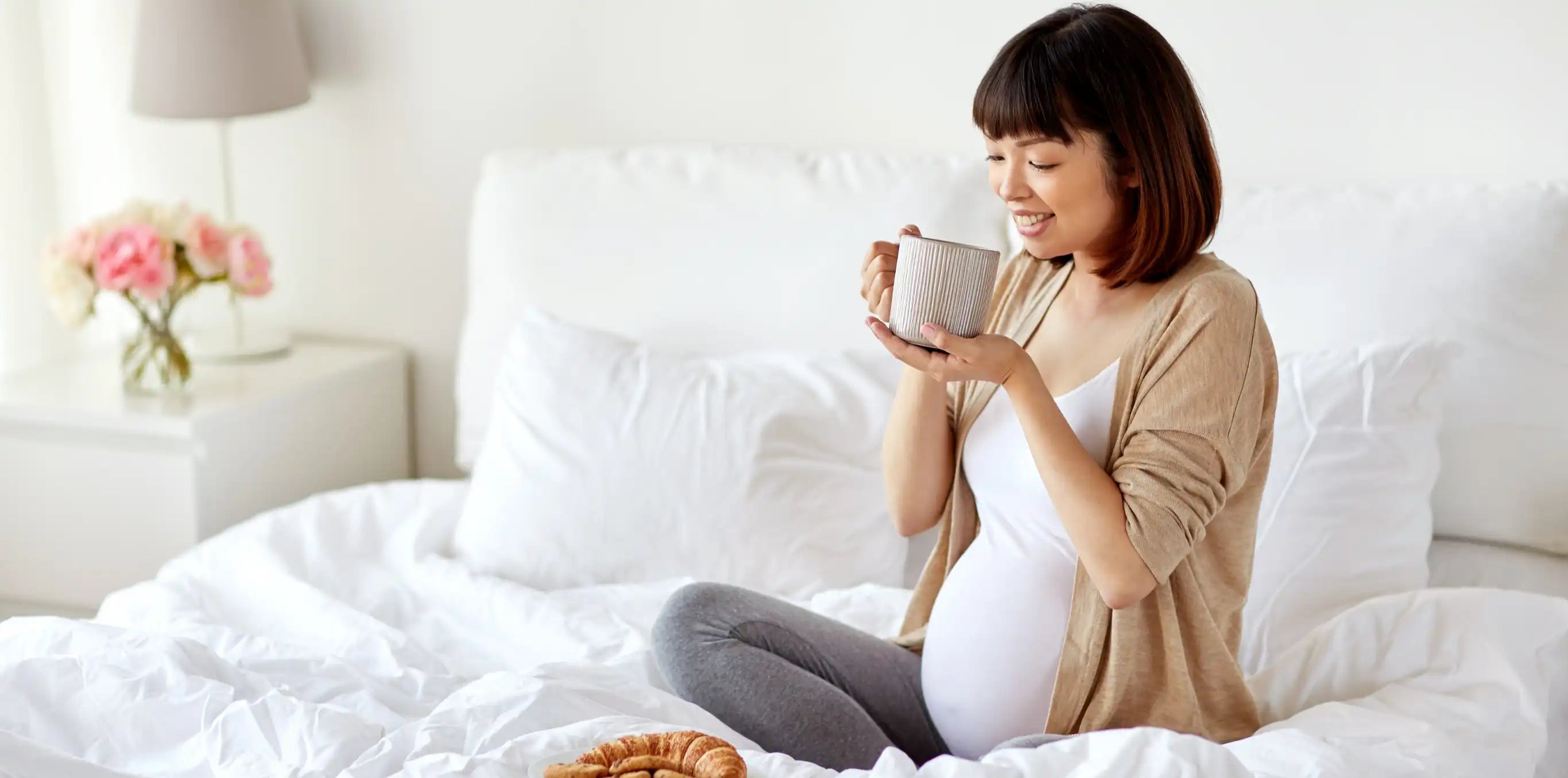 pregnant woman sitting on a bed with a cup of tea and a croissant 