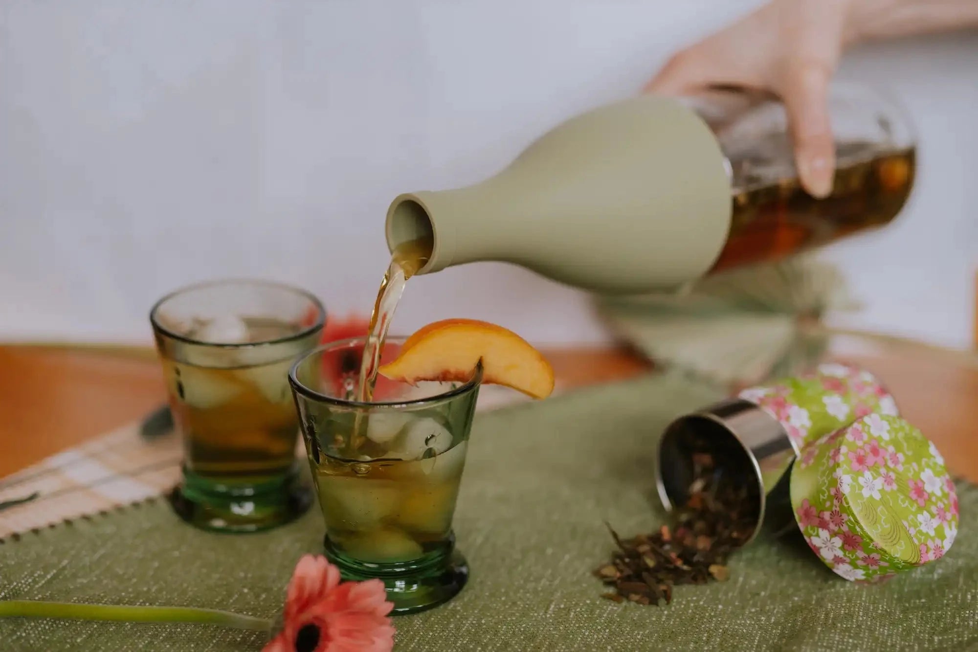 hand pouring iced peach tea into glasses with a peach slice next to a tin of spilled peach tea