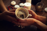 two pairs of hands holding a tea cup with leaves in the bottom and candles in the background