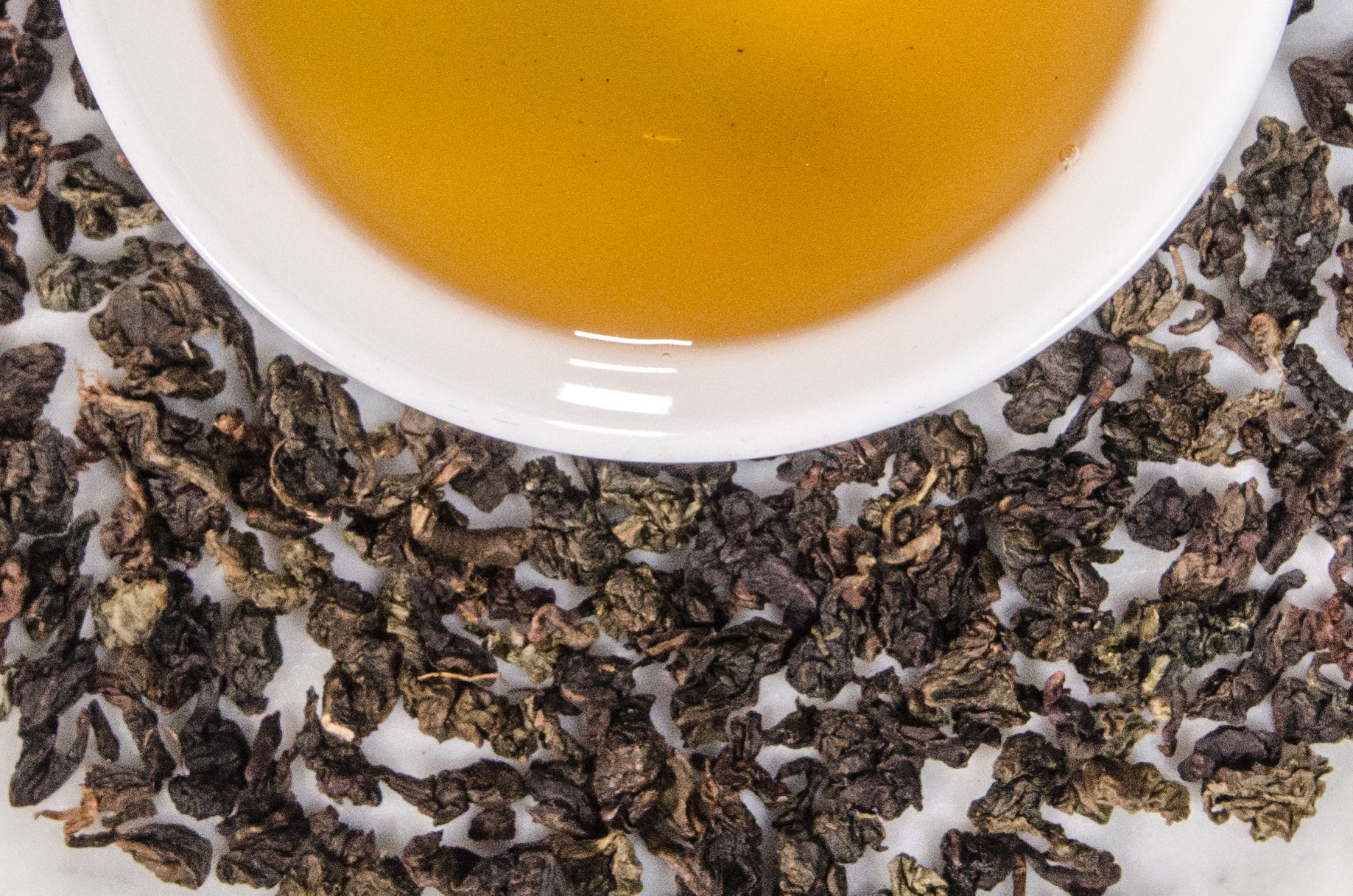 Mucha Tie Guan Yin leaves and brewed tae. Variegated brown and green tightly rolled Taiwanese oolong leaves surround a cup of brewed tawny colored tea.