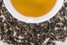 Mucha Tie Guan Yin leaves and brewed tae. Variegated brown and green tightly rolled Taiwanese oolong leaves surround a cup of brewed tawny colored tea.