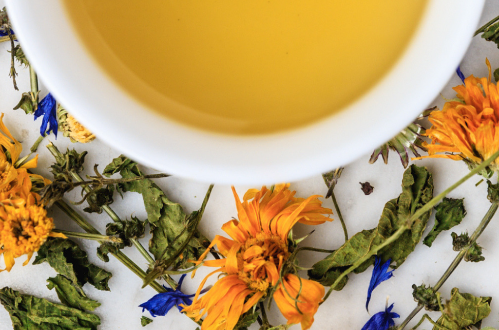 Seasons Tulsi leaves and brewed tea. Green leaves, stems, and bright orange and blue flowers surround a cup of straw-colored brewed herbal tea.