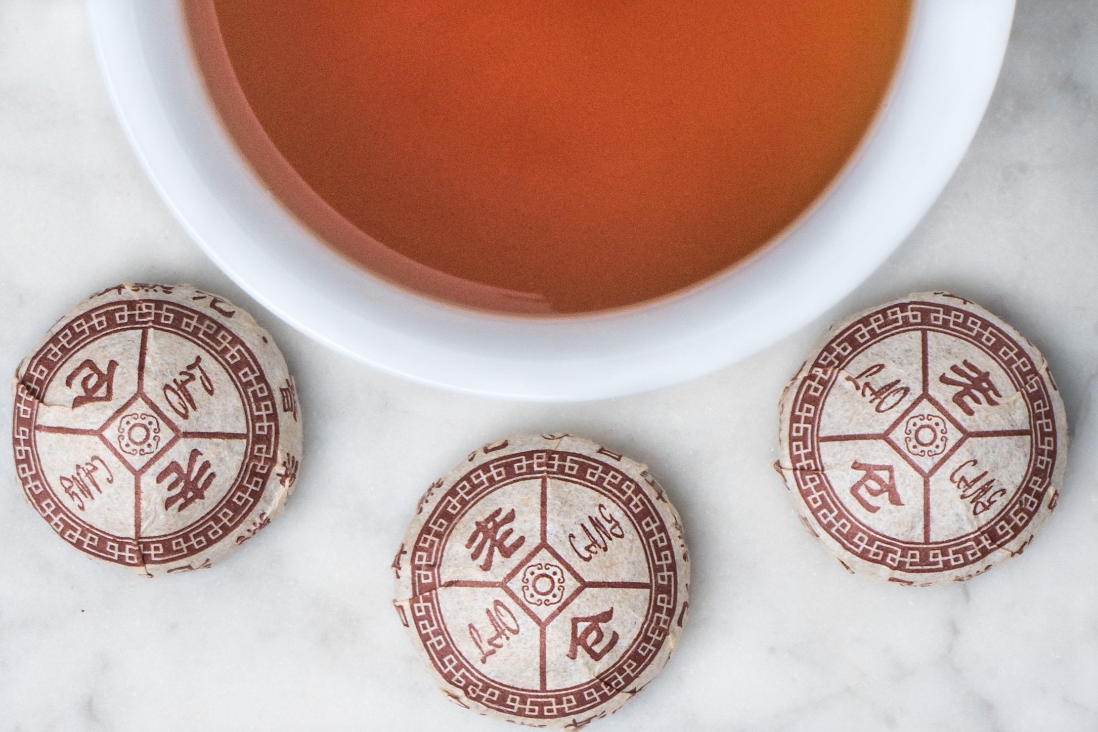 2012 Menghai compressed tea nests and brewed tea. Three small paper wrapped nests of pu erh surround a cup of deep-ochre colored brewed tea.