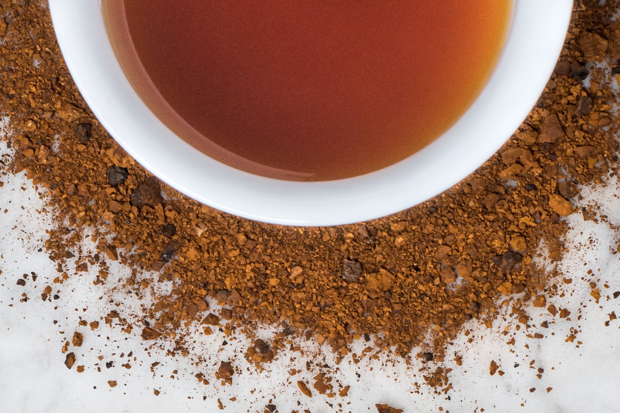 Adirondack Chaga grounds and brewed tea. Coarsely ground, brown chaga mushroom surrounds a cup with dark, reddish-brown brewed chaga. 