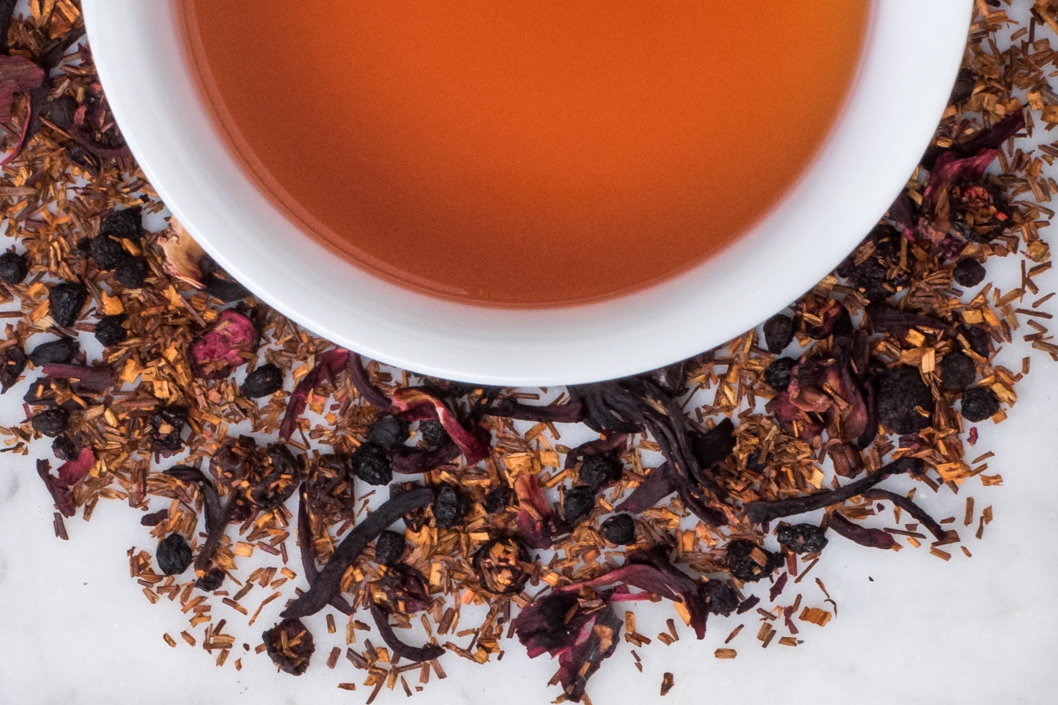 Broadway Berry leaves and brewed tea. Dried blueberry, schisandra berry, hibiscus, and rooibos herbal tea surround a cup of light reddish-brown tea.