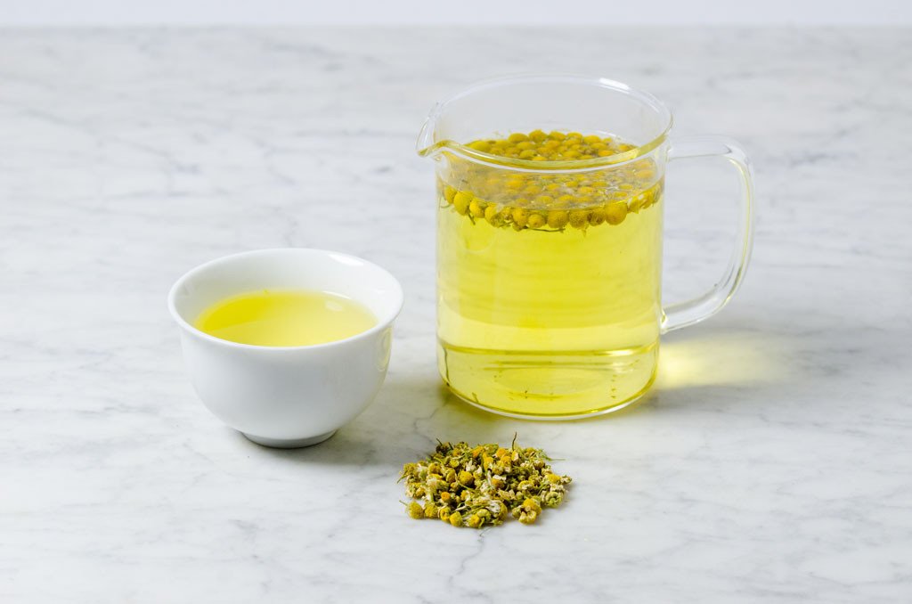chamomile tea in a white tea cup and glass infuser with scattered chamomile flowers