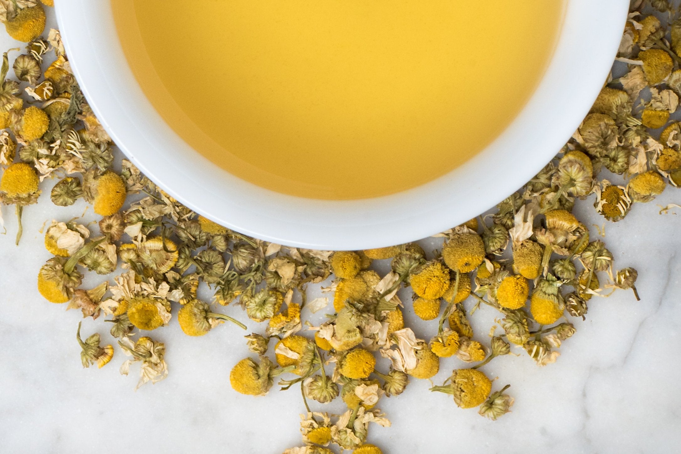 Chamomile Flowers leaves and brewed tea. Plump yellow chamomile buds and petals surround a cup of yellow-hued brewed tea.