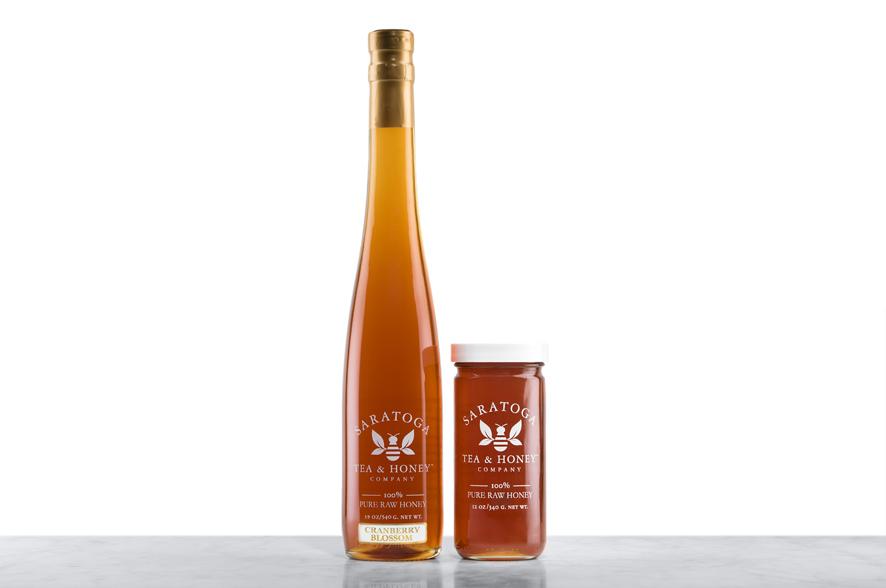 Dark-amber Cranberry Blossom Honey displayed in two glass jars with the logo and the words: "100% Pure Raw Honey". Left: Tall, 19oz bottle shaped like a wine bottle and capped with a cork. Right: 12oz cylindrical bottle with white cap. 