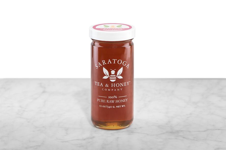 Dark-amber Cranberry Blossom Honey displayed in a 12oz cylindrical glass jar with a white lid. The logo and the words: "100% Pure Raw Honey" are printed on the jar.