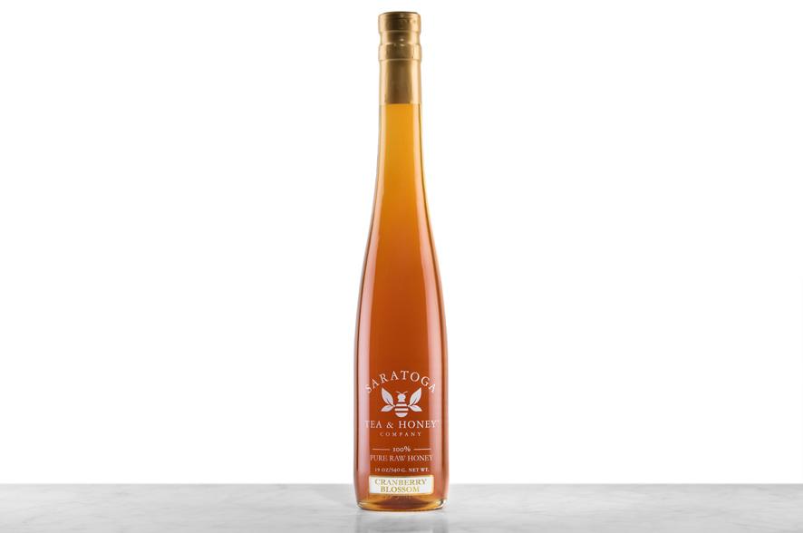 Dark-amber Cranberry Blossom Honey displayed in a tall, 19oz glass bottle shaped like a skinny wine bottle and capped with a cork. Bottle is printed with the logo and the words: "100% Pure Raw Honey".