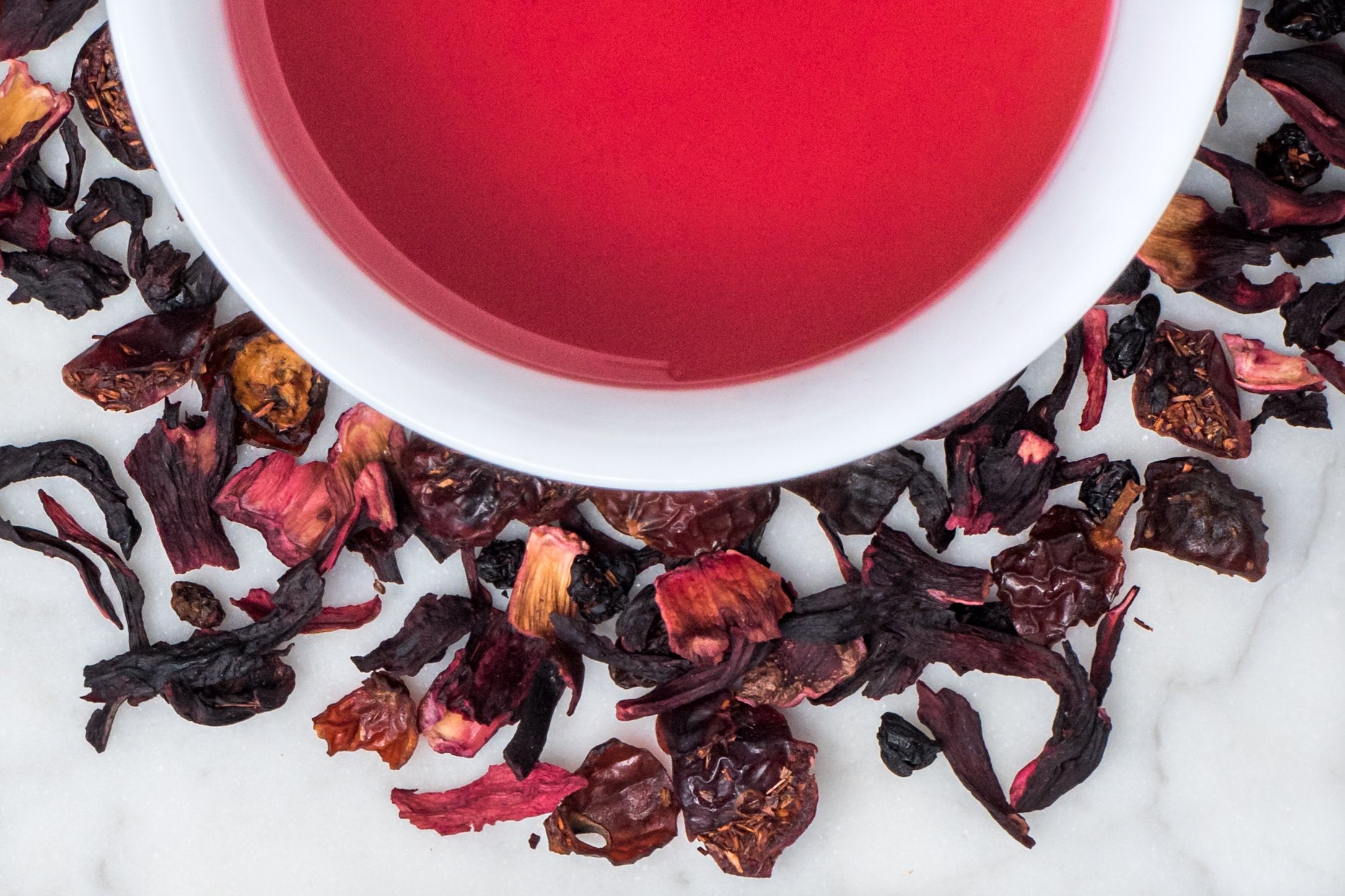 Crimson Berry leaves and brewed tea. Berry-hued dried fruits and hibiscus flowers surround a cup of vibrantly pink brewed tea.