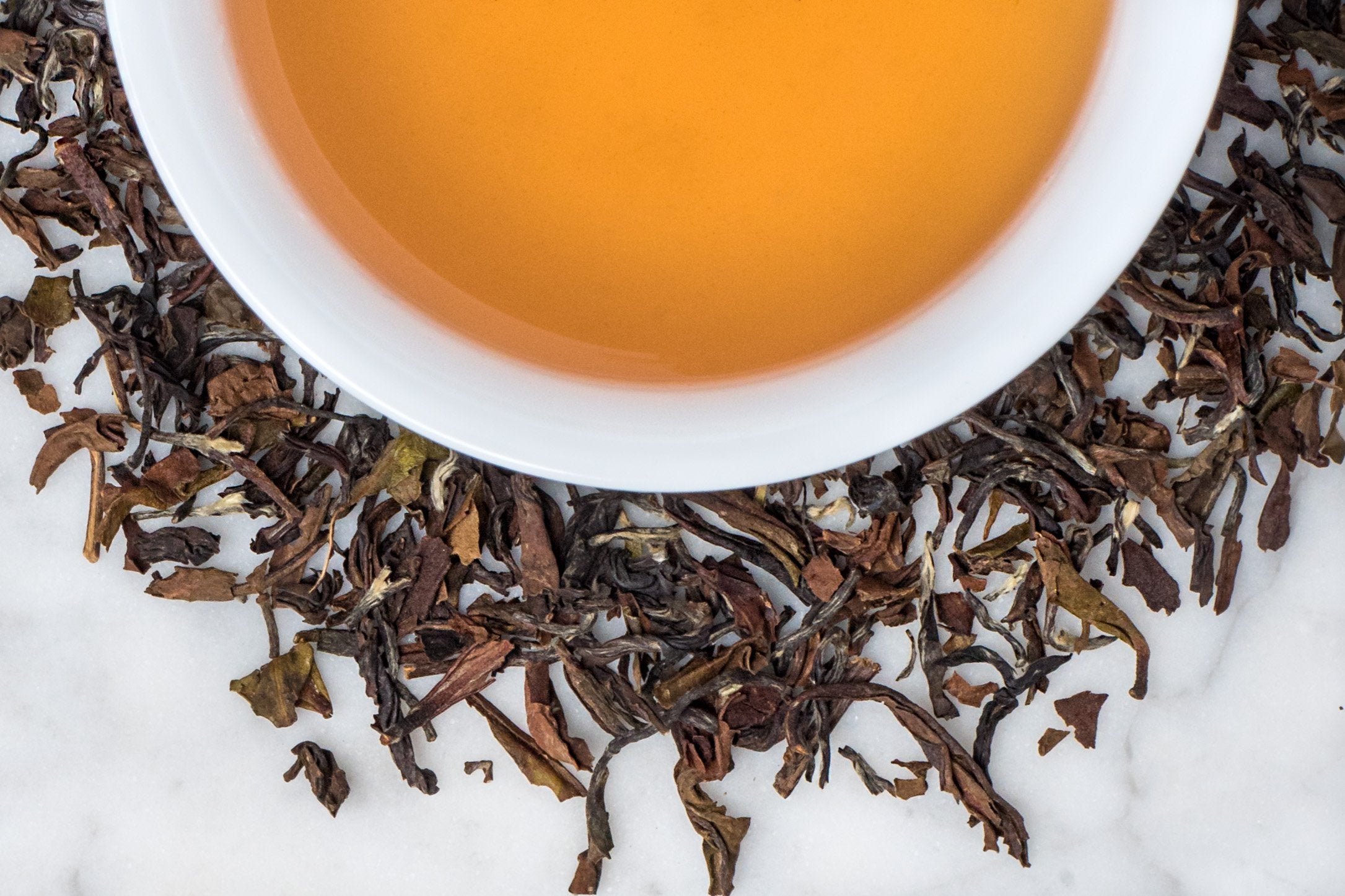 Darjeeling leaves and brewed tea. Twisted brown and khaki leaves surround a cup of golden-brown brewed black tea.