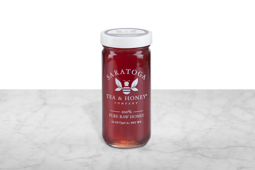 Reddish-amber Elderberry Infused Honey displayed in a 12oz cylindrical glass jar with a white lid. The logo and the words: "100% Pure Raw Honey" are printed on the jar.