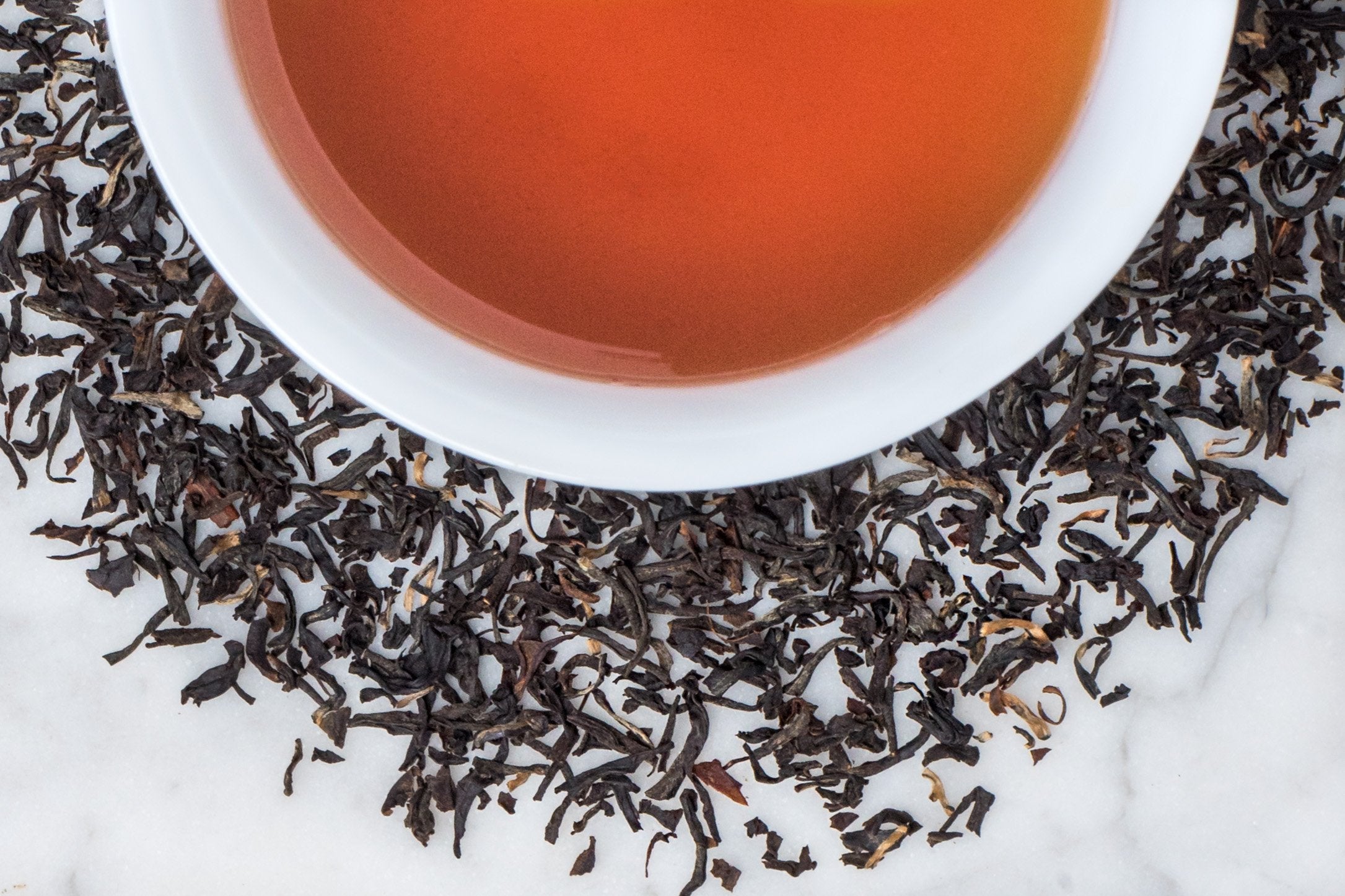 English Breakfast Assam Banaspaty leaves and brewed tea. Small but full twisted black leaves and golden buds surround a cup of russet-hued brewed tea.