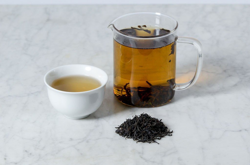 golden black tea infusion presented in a white cup next to a glass infuser with a pile of loose tea leaves