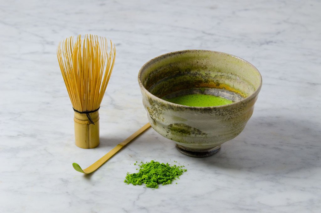 matcha chasen, bamboo matcha scoop, matcha powder, and chawan (bowl) of matcha next to each other on a marble background