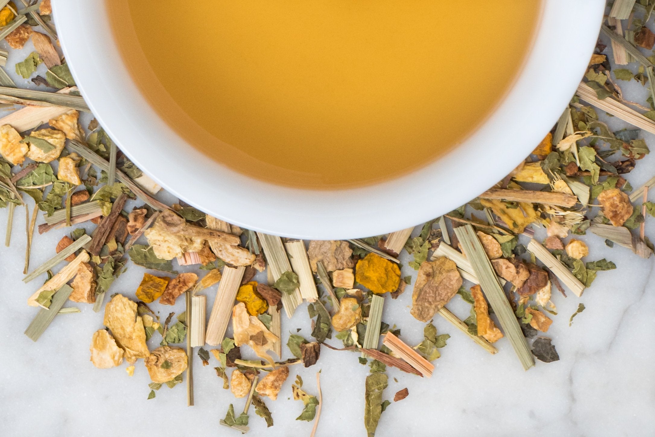 Flu Shot leaves and brewed tea. An earthy mix of dried lemongrass, lemon verbena, turmeric and other spices surround a cup of dark yellow brewed tea.