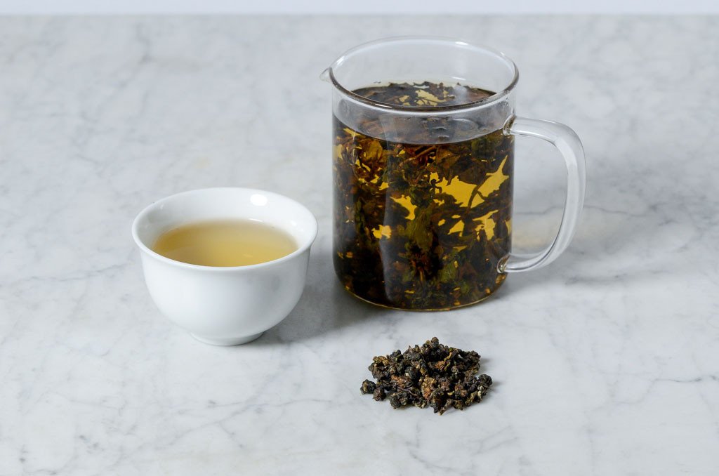 Guei Fei bug bitten oolong steeped in a glass infuser and served in a white cup alongside rolled oolong leaves