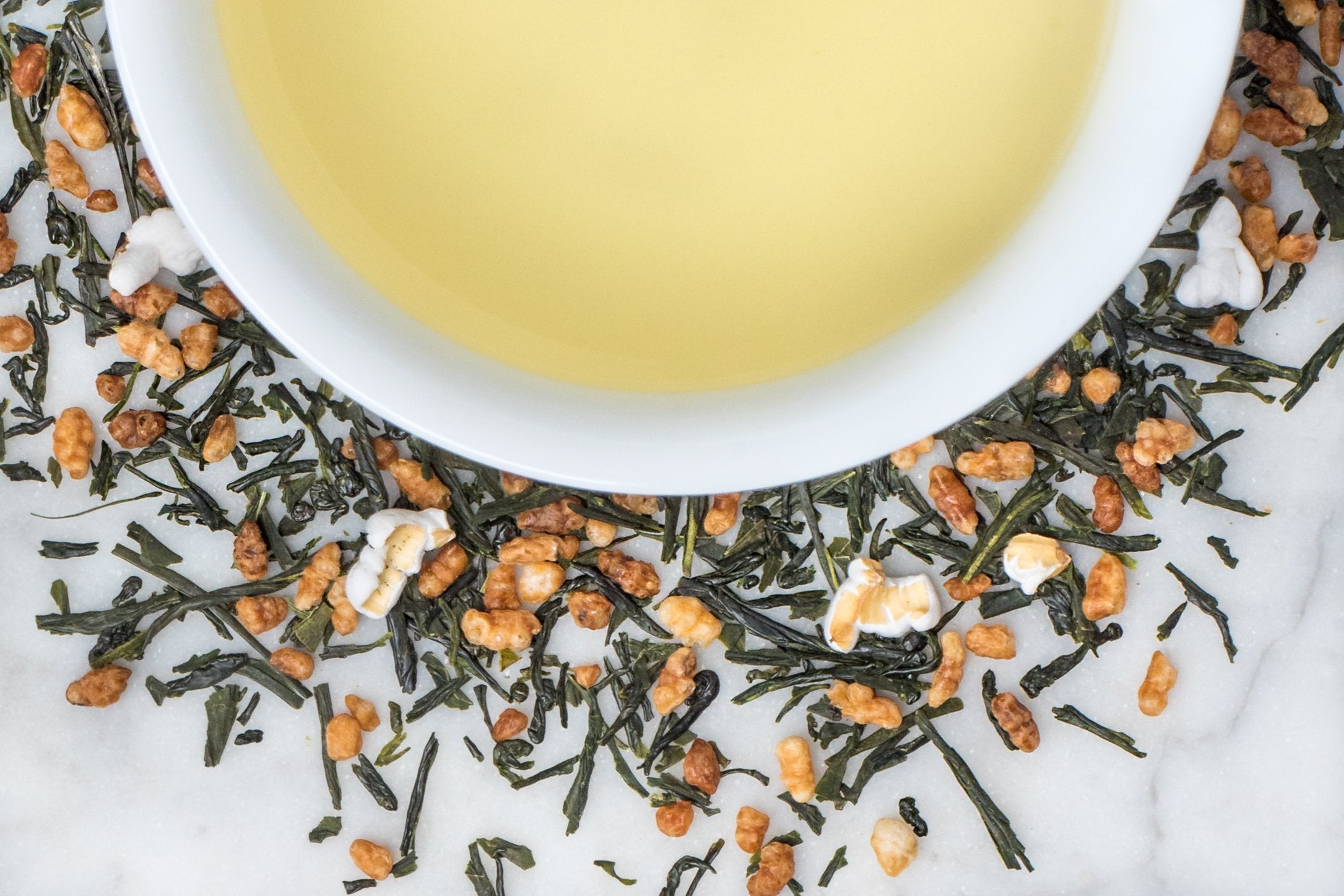 Genmai Cha leaves and brewed tea. Small, dark green leaves and brown popped rice surround a cup of palest pastel-yellow brewed tea.