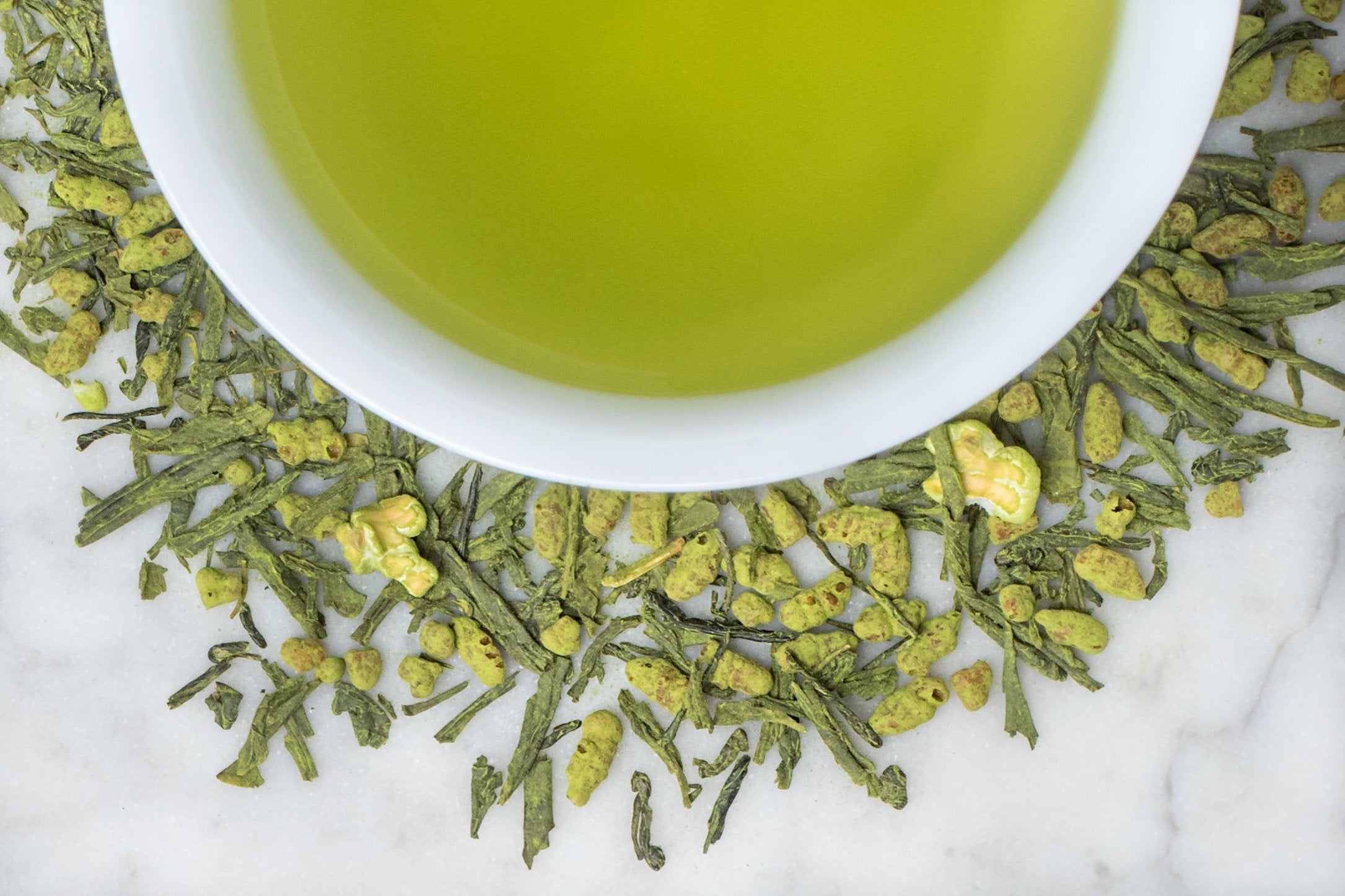 Genmai Cha Sencha Matcha leaves and brewed tea. Small, bright green tea leaves and pieces of popped rice dusted with green matcha surround a cup of grass-green brewed tea.