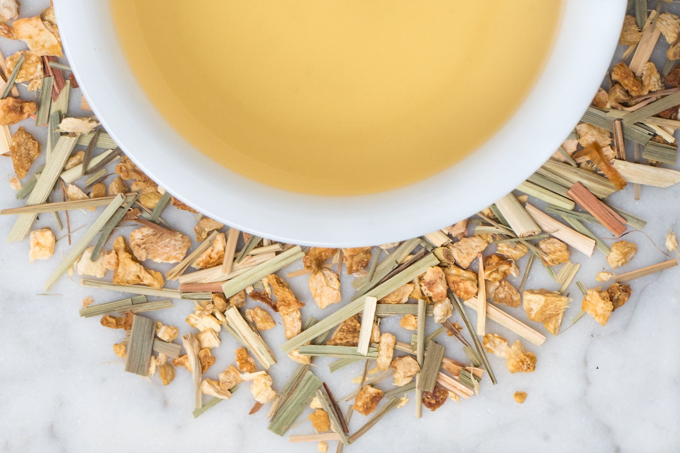 Ginger Lemon Zen leaves and brewed tea. Torn lemongrass, ginger, and lemon peel surround a cup of light yellow brewed tea.