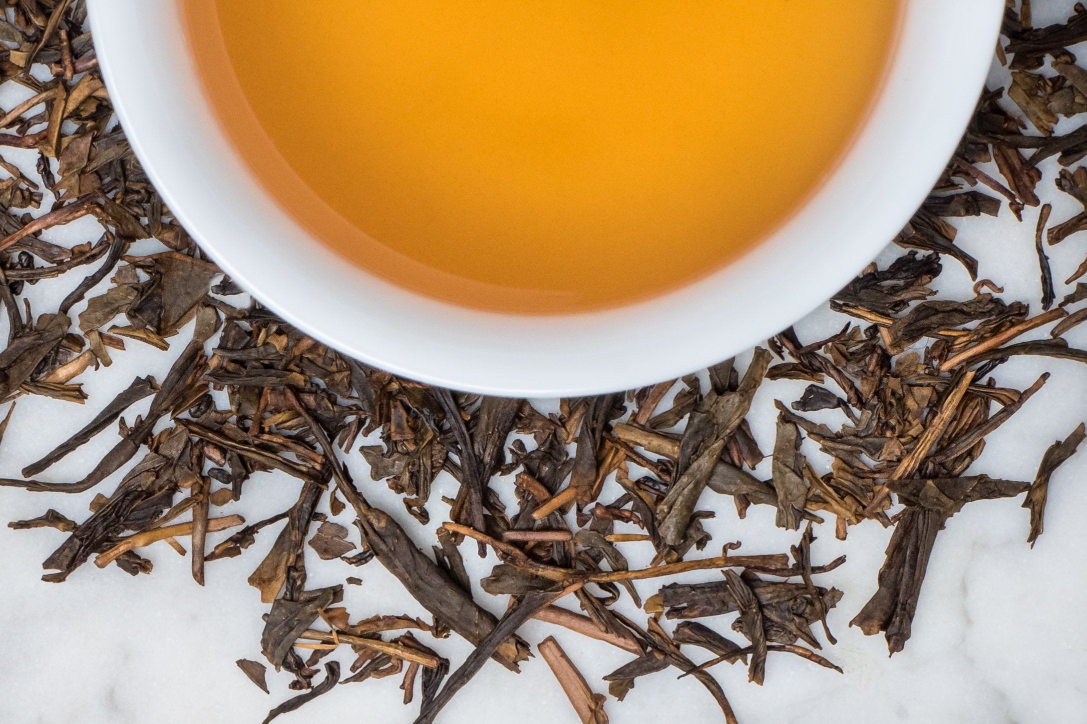 Hojicha leaves and brewed tea. Dark and medium-brown leaves and stem pieces surround a cup of golden-amber brewed tea.