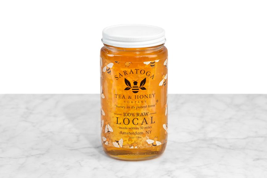 Golden Local Wildflower Honeycomb soaked in honey and displayed in a 16 oz cylindrical glass jar with a white lid. The logo and the words: "100% Raw Local, Amsterdam NY" are printed on the jar.