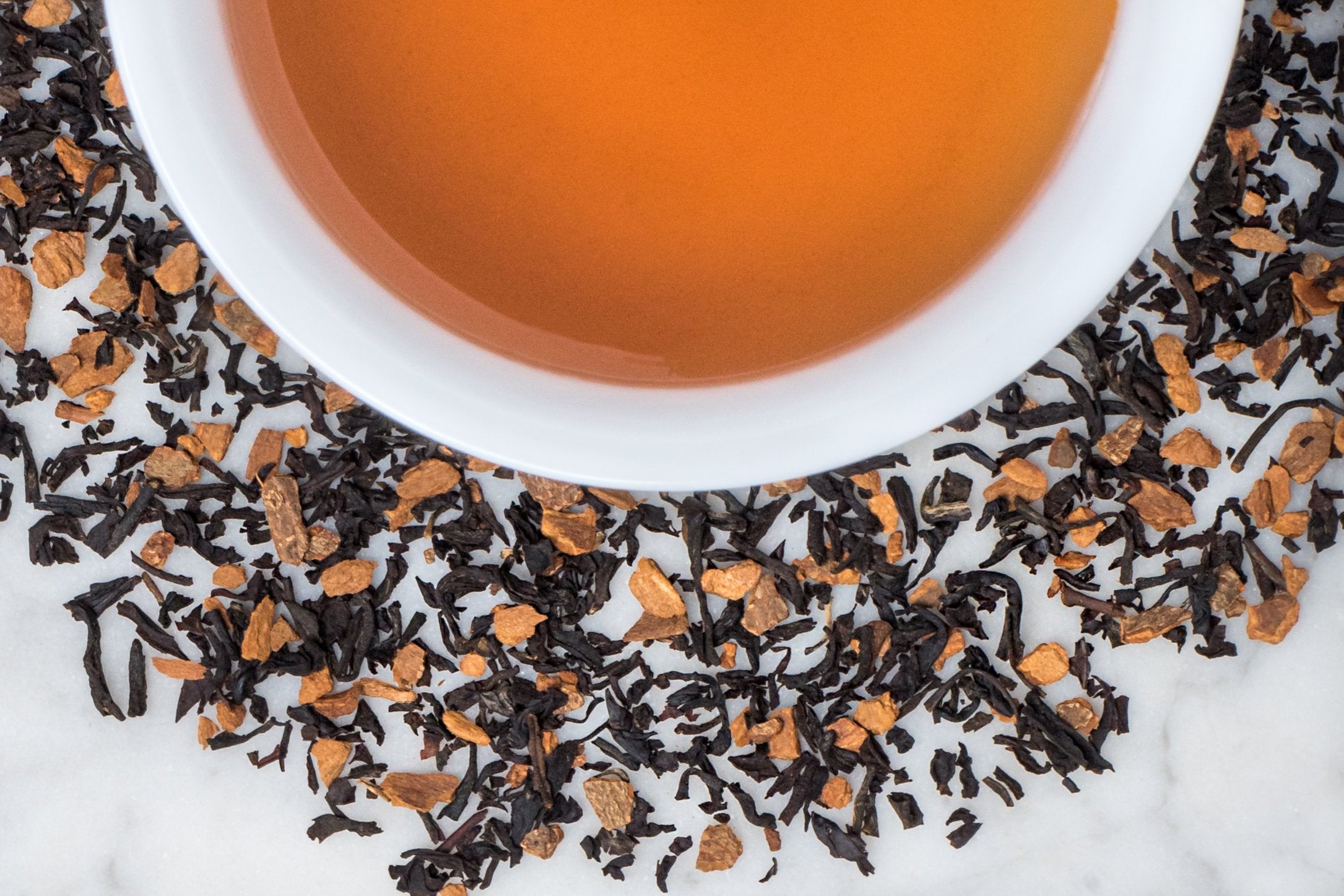 Istanbul Apple leaves and brewed tea. Short black tea leaves and cinnamon pieces surround a cup of dark-amber-colored brewed tea.