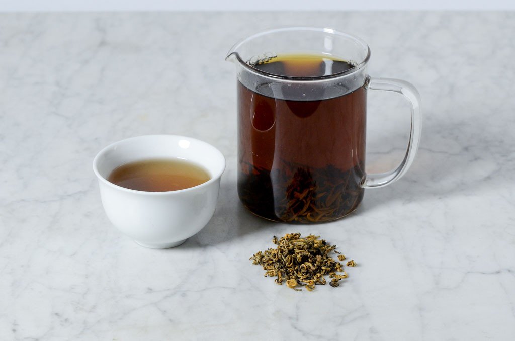 Jin Die brewed and presented in a white cup beside a glass infuser and pile of rolled golden bud Jin Die black tea