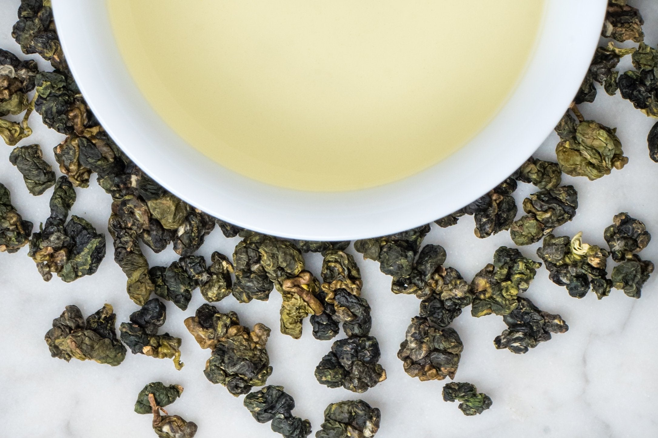 Jin Xuan 'Milk' Oolong leaves and brewed tea. Tightly rolled, imperfect balls of leaves in varying green hues surround a cup of very pale pastel yellow brewed tea.