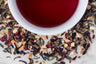 Joyful Joyful holiday tea blend leaves and brewed tea. Colorful green and black tea leaves, hibiscus flowers, and spices surround a cup of crimson-hued brewed tea.