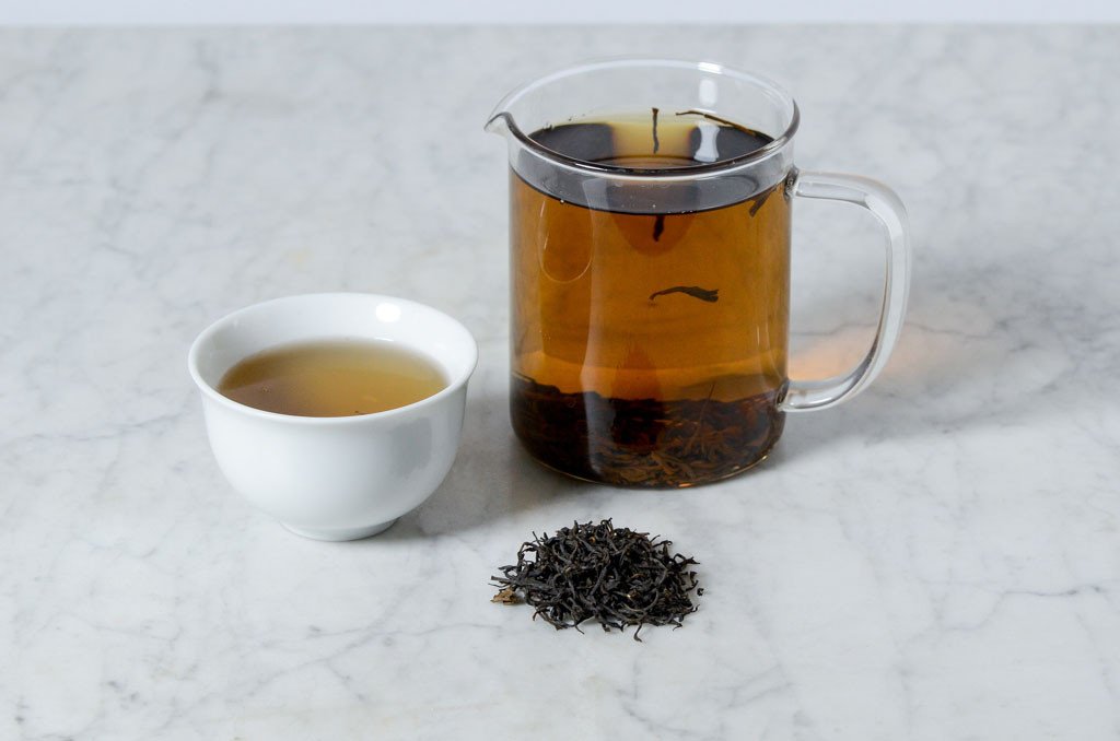 infused black tea in a white cup beside a glass infuser presented with a pile of black tea leaves on a marble background
