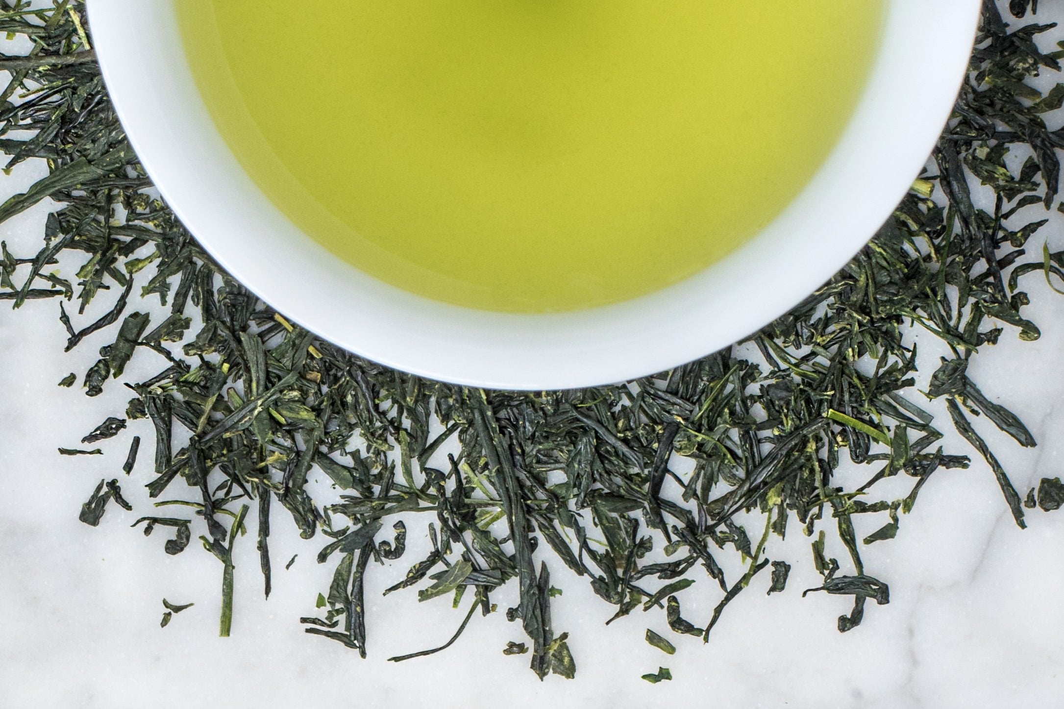 Kabusecha Takamado green tea leaves and brewed tea. Small and thin, needle-like dark green leaves surround a cup of lime-green brewed tea. 