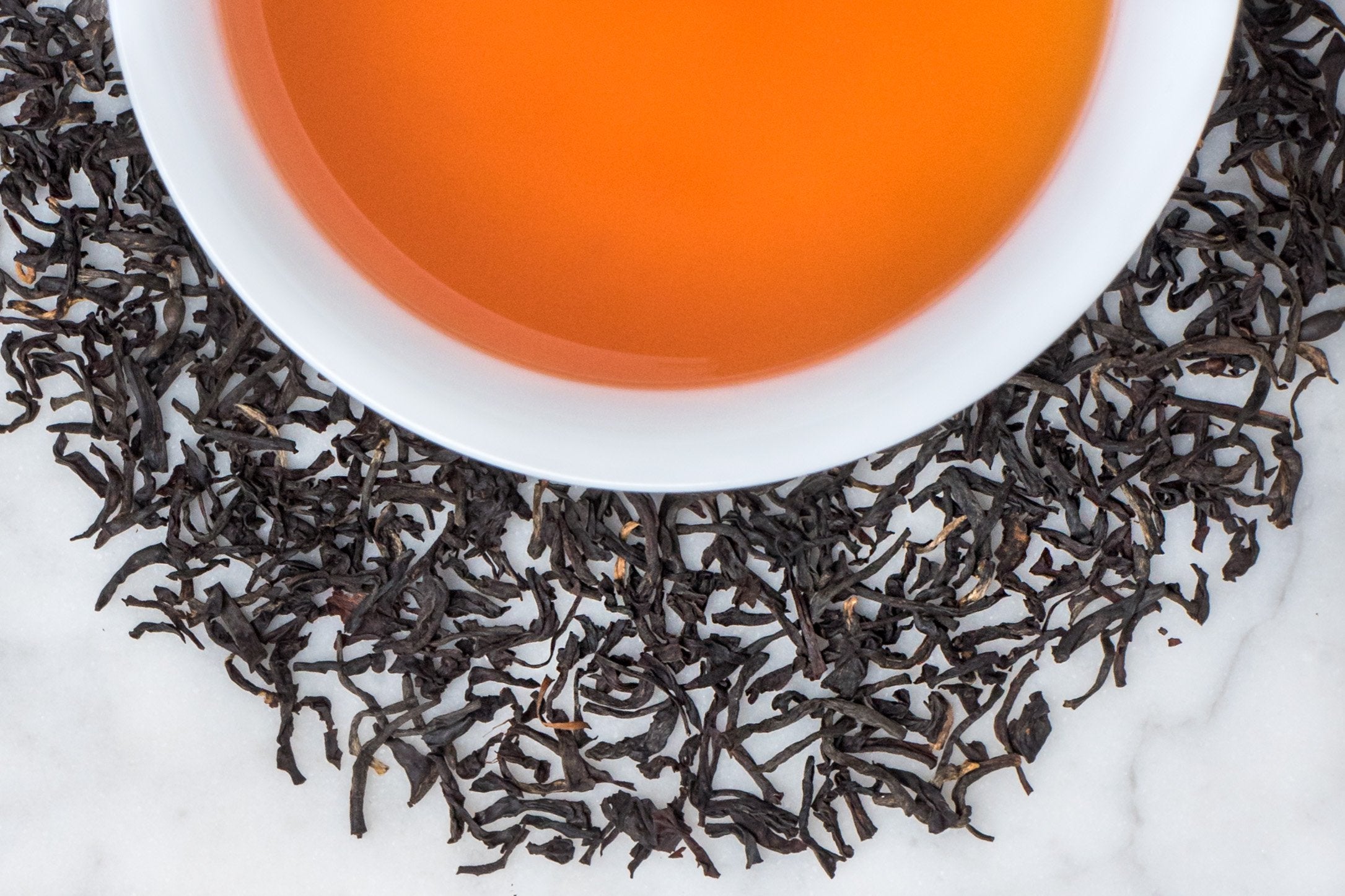 Kenya Kangaita leaves and brewed tea. Small, twisted black tea leaves surround a cup of orangey-brown brewed black tea.