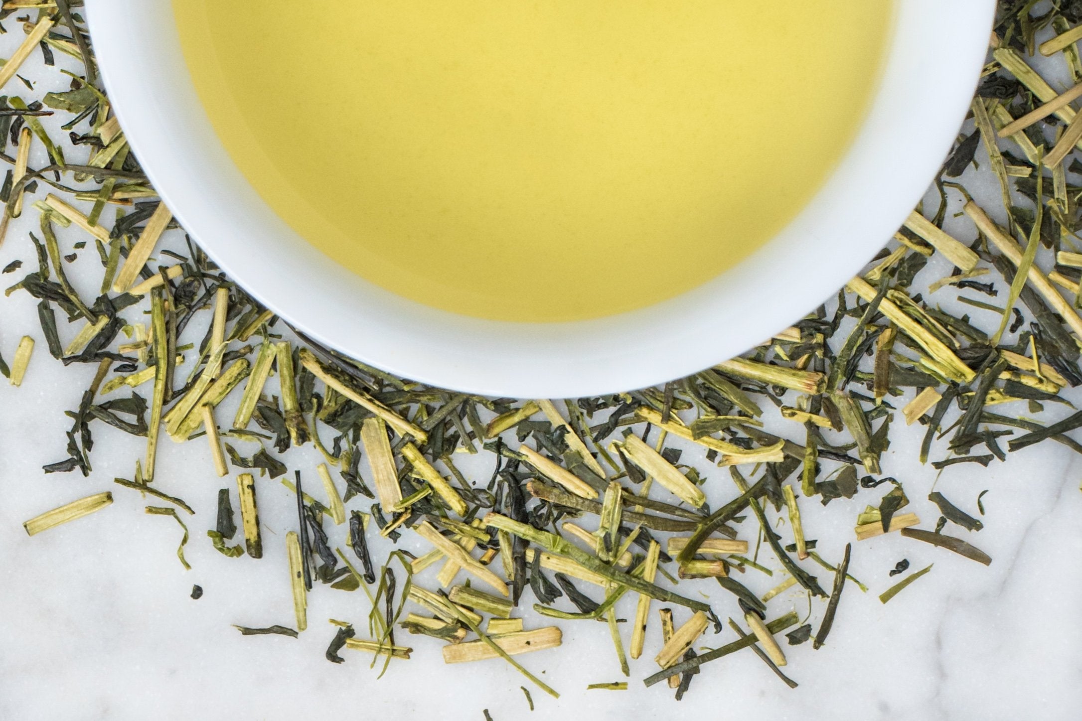 Kukicha leaves and brewed tea. Short, thin, stick-like leaves of light and dark green tea surround a cup of bright yellow-green brewed tea.