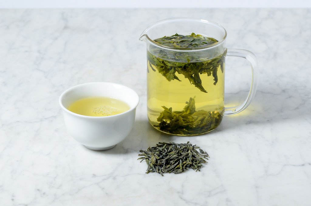 loose leaf lu an gua pian green tea in a pile in front of a brewing glass pitcher of green tea and a white cup of green tea