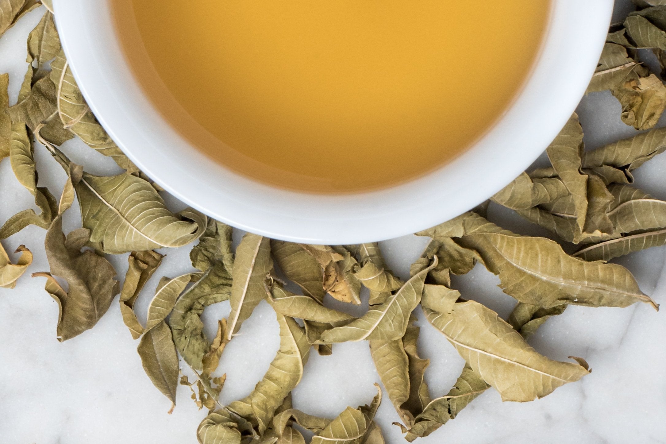 Lemon Verbena leaves and brewed tea. Large, full, khaki green dried lemon verbena leaves surround a cup of golden-yellow brewed tea.