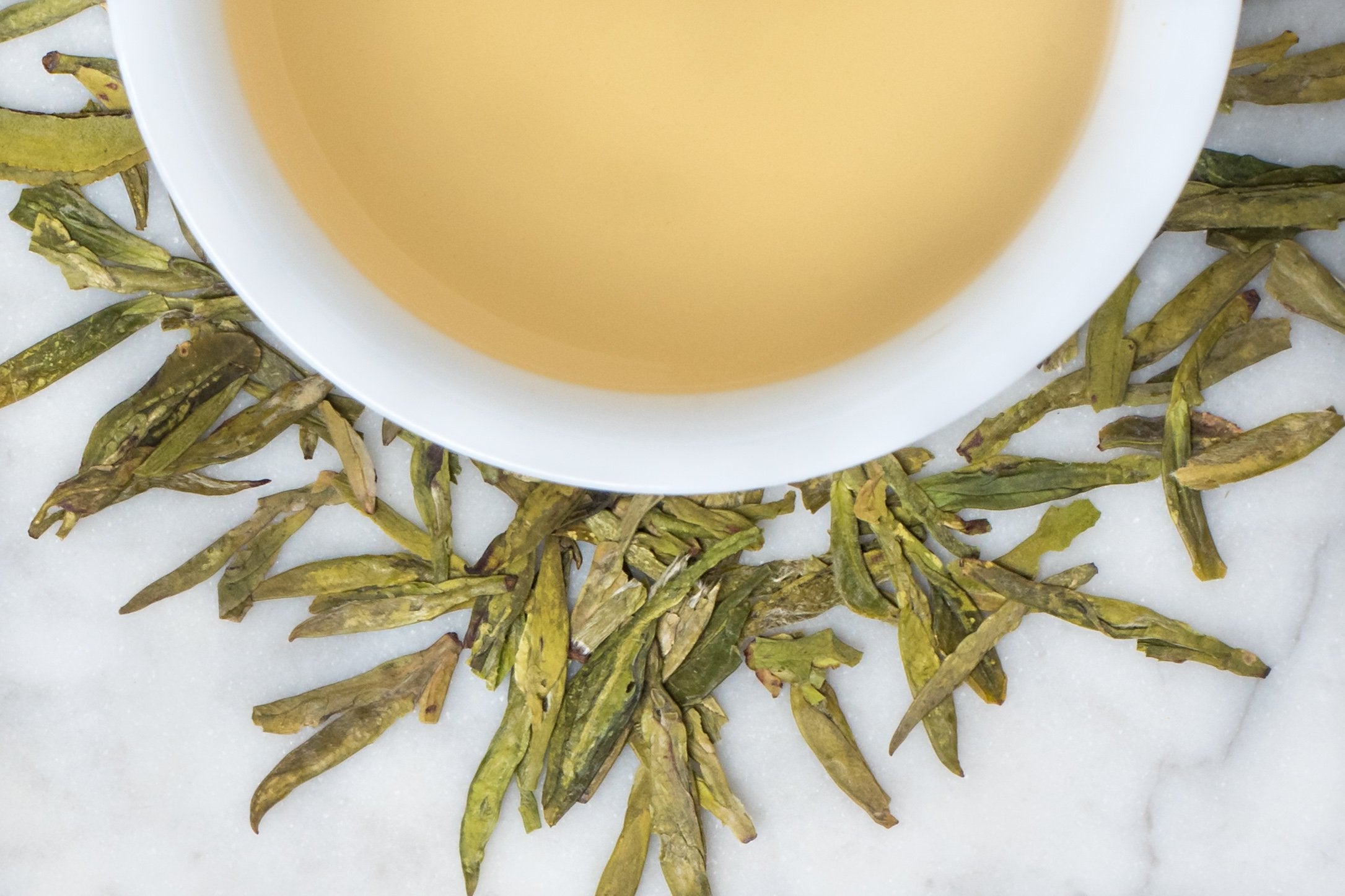 Long Jing Zhejiang (Dragonwell) leaves and brewed tea. Long and flat light green tea leaves surround a cup of palest peachy yellow brewed tea.
